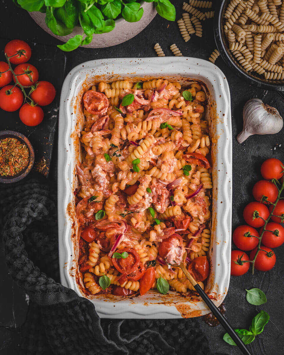 Das fertige Baked Feta Pasta Rezept: Nudeln mit gebackenem Schafskäse, Tomaten und frischen Kräutern in einer Auflaufform auf dunklem Untergrund
