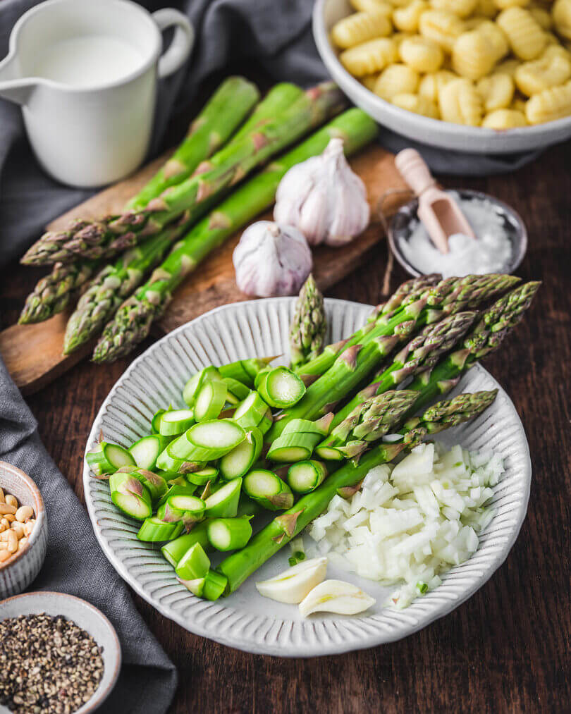 Frisch vorbereitete Zutaten für Gnocchi mit grünem Spargel: geschnittener Spargel, gehackte Zwiebeln, Knoblauch, Gnocchi und Sahne auf einem Holztisch.
