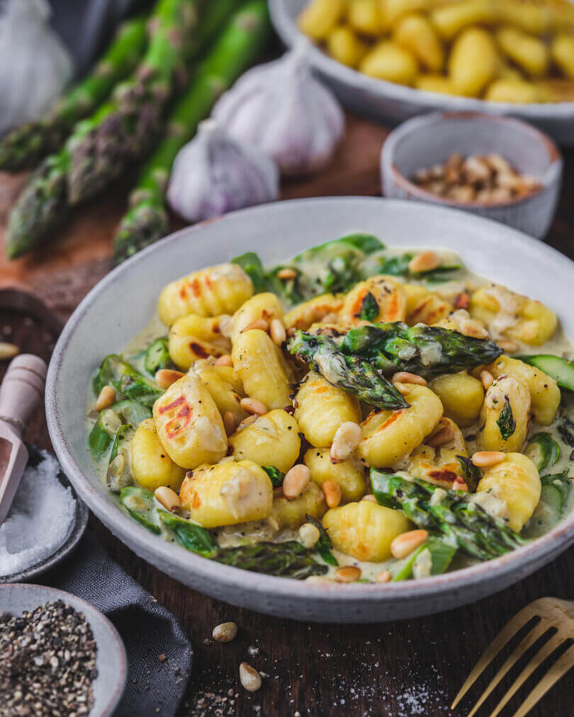 Servierfertige Gnocchi mit grünem Spargel und Sahnesauce, bestreut mit gerösteten Pinienkernen und frisch gemahlenem Pfeffer.