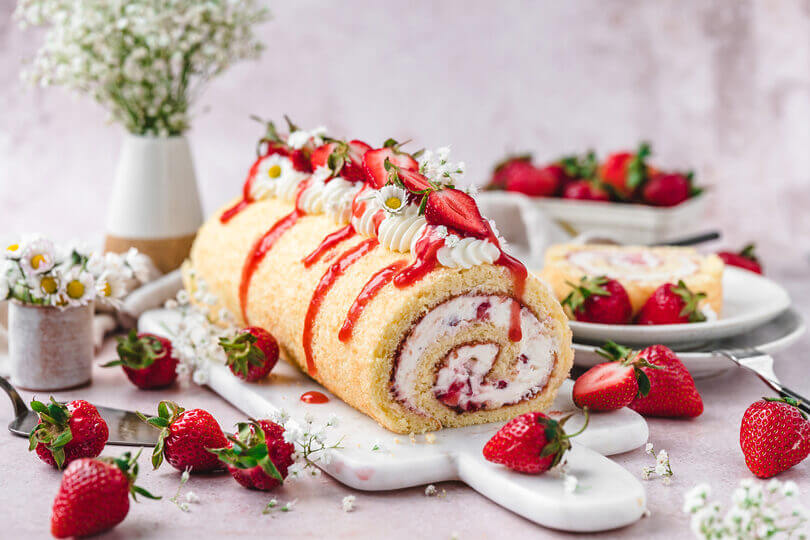 Ganze Erdbeer-Biskuitroulade auf einer Marmorplatte, dekoriert mit frischen Erdbeeren und Sahne.