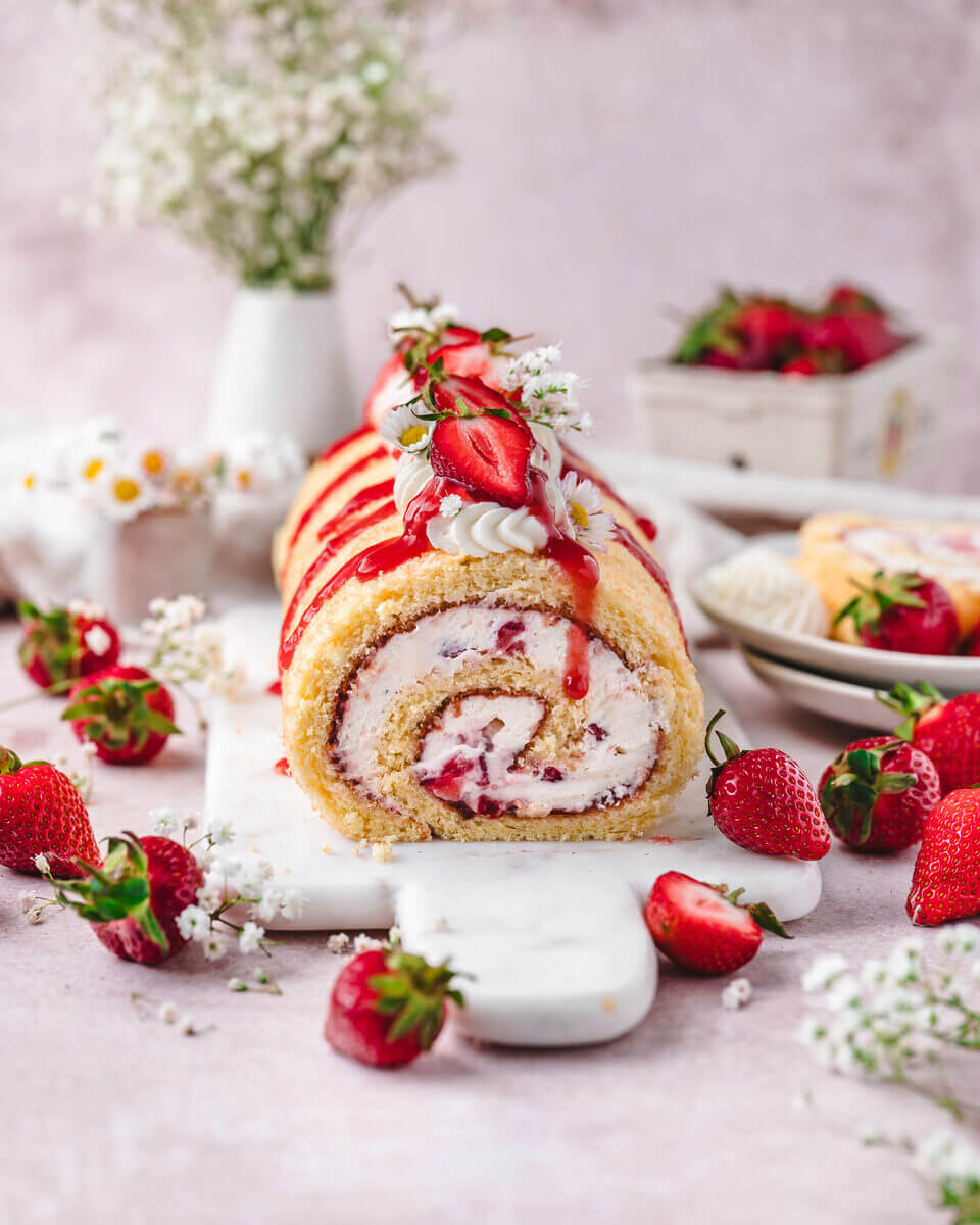Frontale Ansicht einer sommerlichen Erdbeerroulade mit Sahne, Konfitüre und Erdbeerdeko angerichtet auf einer Marmorplatte.