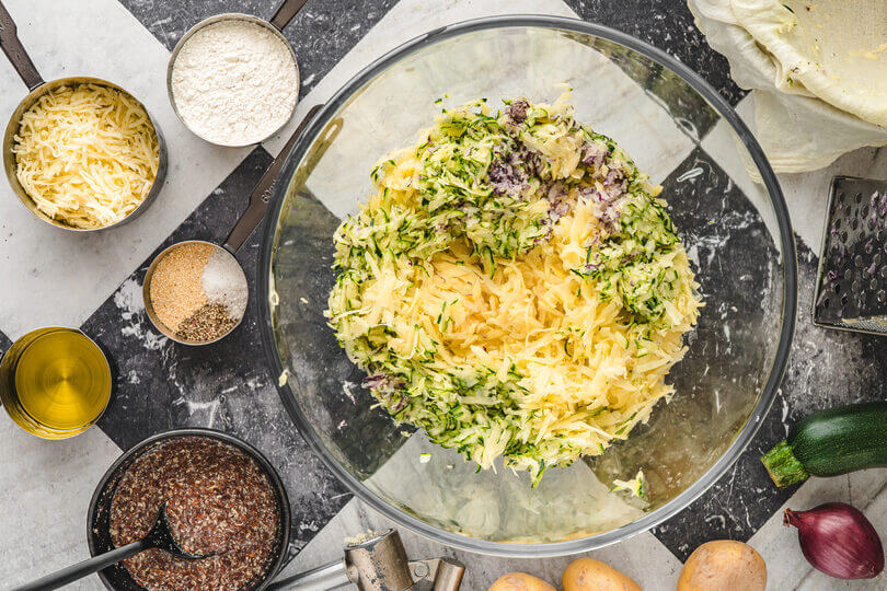 Alle Zutaten für die Gemüserolle in kleinen Schüsseln: geraspelte Zucchini, Kartoffeln, rote Zwiebel, Leinsamen, Käse, Mehl, Öl und Gewürze.