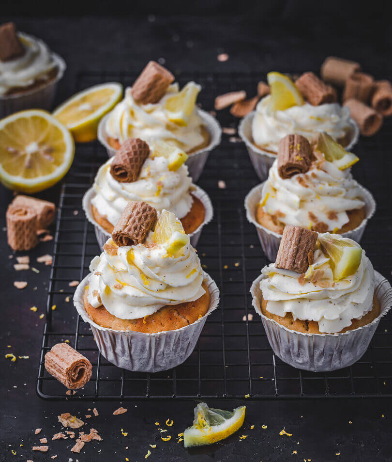 Frisch gebackene Zitronenmuffins mit cremigem Frosting, dekoriert mit Zitronenscheiben und Waffelröllchen auf einem Kuchengitter