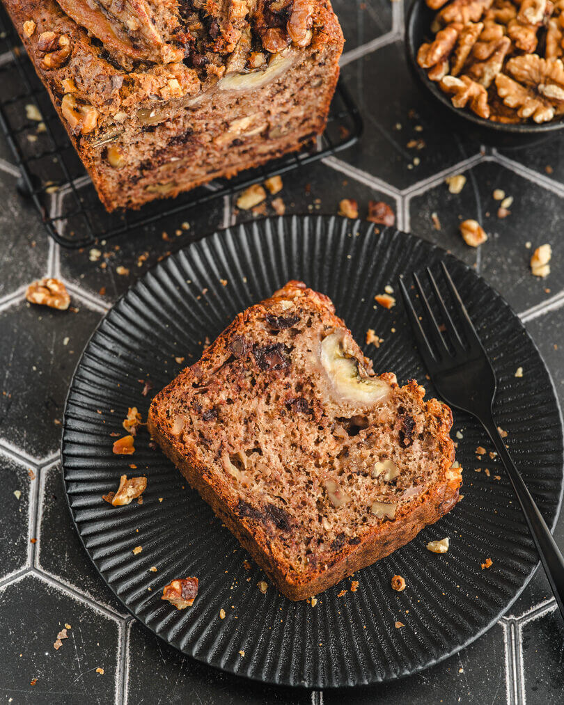 Ein saftiges Stück Bananenbrot mit Walnüssen und dunkler Schokolade angerichtet auf einem schwarzem Teller